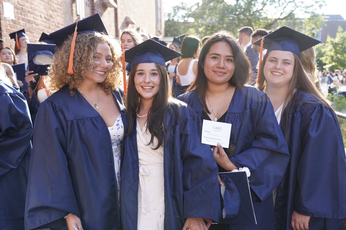Four smiling graduates in navy blue gowns and caps with orange tassels pose at an outdoor commencement.