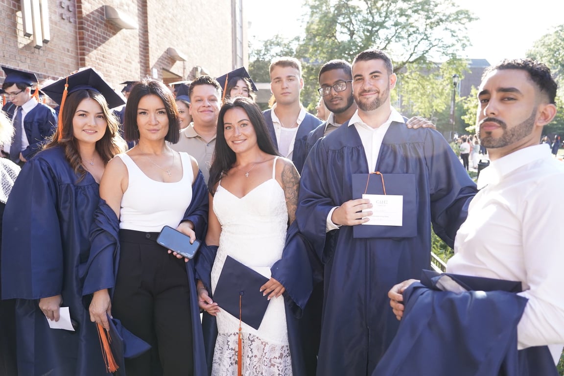 CAHE graduates and guests pose outdoors near a brick building; navy gowns and a CAHE certificate are visible.