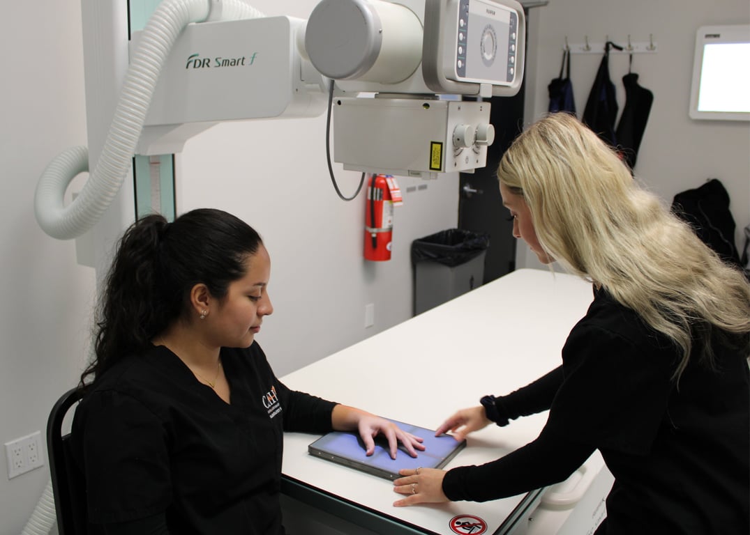 A radiography student in black scrubs positions a classmate’s hand on a digital X-ray receptor under a Fujifilm X-ray tube.