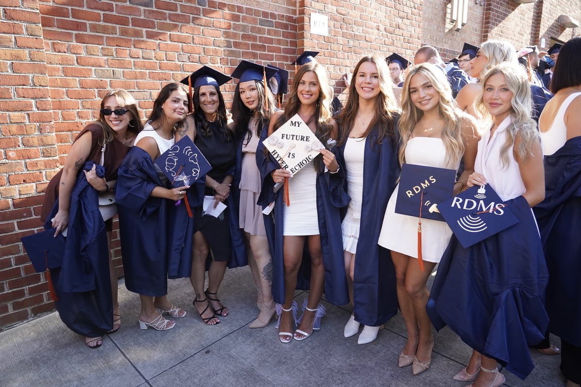 Diagnostic medical sonography graduates in navy gowns pose with RDMS-decorated caps; one cap reads “My Future is Hyperechoic.”