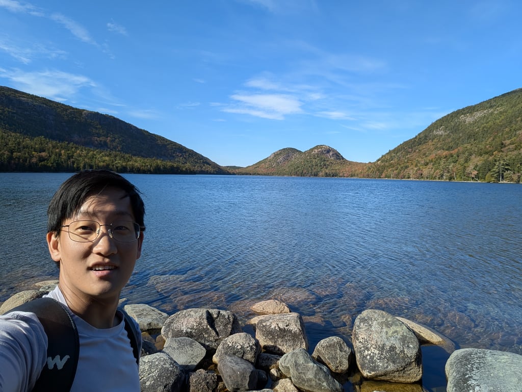 Image with caption 'A beautiful little lake/large pond with some rolling hills in the distance. One side of the pond is a traditional trail running alongside rocks; the other side is mostly boardwalk due to some revegetation work ongoing.'