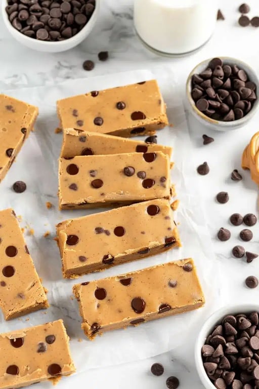 Sliced cookie dough protein bars arranged on parchment with chocolate chips and peanut butter
