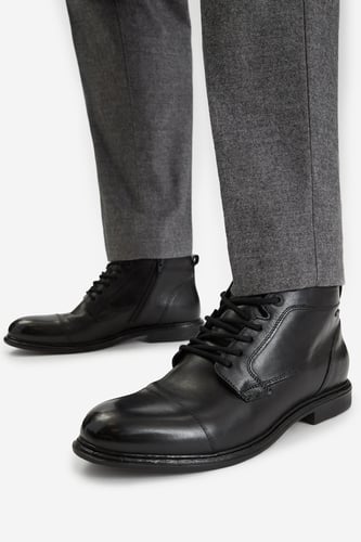 Model w spodniach prezentuje sznurowane buty za kostkę, ujęcie z przodu.