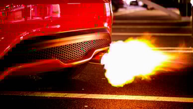 Detail zadní části zářivě červeného sportovního vozu s dramatickým plamenem šlehajícím z výfukové trubky. Tento dynamický snímek zdůrazňuje výfukový systém vozidla, vizuálně reprezentuje výkon a potenciál pro neobvyklé emise. Ideální pro články pojednávající o problémech s výfukem, ladění výkonu nebo důležitosti správně fungujícího výfukového systému pro zdraví motoru a kontrolu emisí.