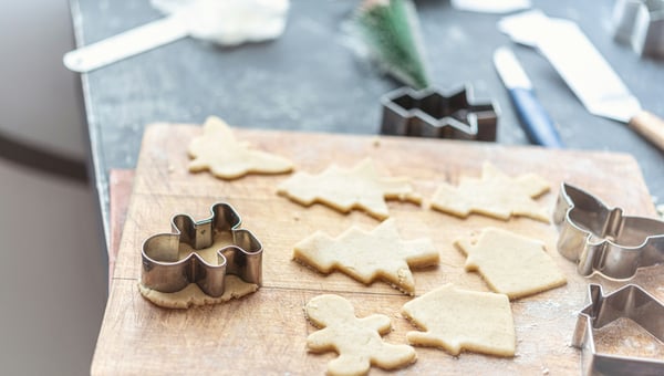 Detailní pohled na dřevěné prkénko s vykrojeným syrovým těstem na cukroví, obklopené kovovými formičkami různých tvarů, jako jsou stromečky a perníčci. Zdůrazňuje použití 3D tiskáren pro výrobu bezpečných kuchyňských pomůcek.