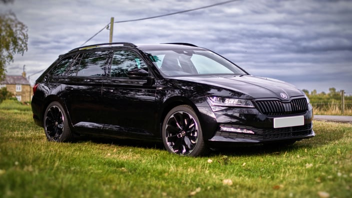 Černá Škoda Superb Combi III facelift s černými litými koly zaparkovaná na trávě pod mrakem v přírodě.