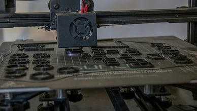 Ruce v rukavicích brousí 3D tištěný model, v pozadí jsou vidět nástroje pro post-processing jako štětce, barvy a tmely. Ilustruje proces dokončovacích prací na 3D výtisku.