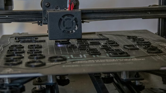 Ruce v rukavicích brousí 3D tištěný model, v pozadí jsou vidět nástroje pro post-processing jako štětce, barvy a tmely. Ilustruje proces dokončovacích prací na 3D výtisku.