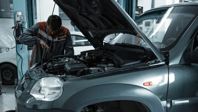 Автомеханик в рабочей форме наклоняется над открытым капотом темного внедорожника и проводит диагностику двигателя в чистом автосервисе с другой машиной на заднем плане.