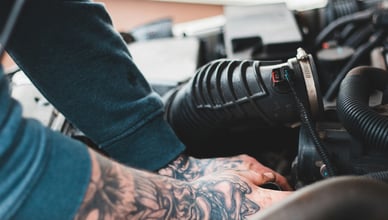 Механик с татуировками на руках работает в моторном отсеке автомобиля, выполняет обслуживание и чистку топливных форсунок.