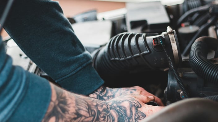 Механик с татуировками на руках работает в моторном отсеке автомобиля, выполняет обслуживание и чистку топливных форсунок.