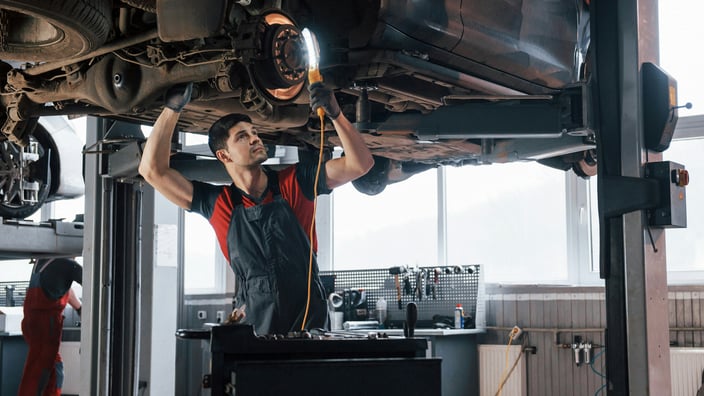 Механик в сервисе проводит диагностику подвески, с фонарем проверяет стойки стабилизатора и ось на поднятом автомобиле.