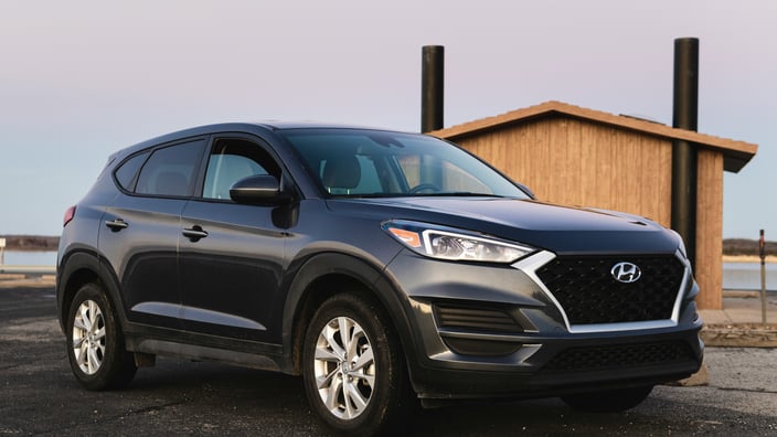 Tmavě šedé SUV Hyundai Tucson zaparkované na asfaltové ploše u dřevěné budovy během západu slunce.
