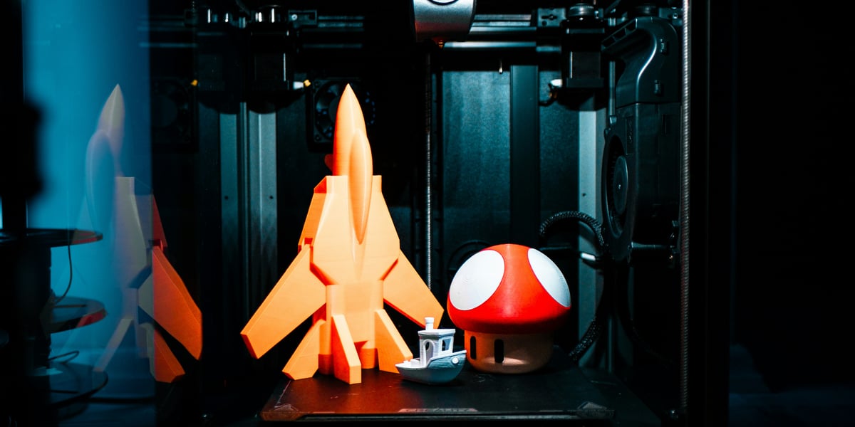 3D tiskárna v temné místnosti osvětlená modrým a červeným světlem. Uvnitř tiskárny leží na podložce tři 3D tištěné modely: velká oranžová stíhačka nebo raketa, malý bílý model lodi a velká červenobílá houba ve stylu Super Mario. V popředí je vidět ovládací dotykový displej.