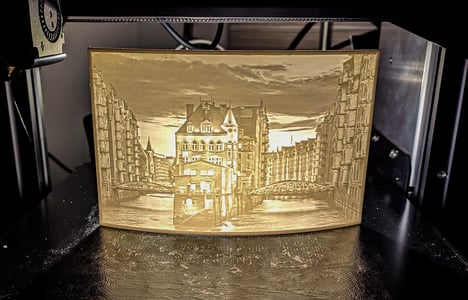 3D tiskárna vytváří světle zbarvený, průsvitný obraz města s řekou a budovami. Objekt je typickým příkladem litofánie, což je 3D vytištěná fotografie, která září při osvětlení a je ideálním personalizovaným dárkem.