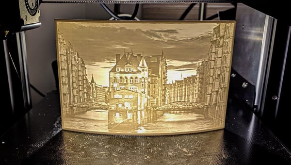 3D tiskárna vytváří světle zbarvený, průsvitný obraz města s řekou a budovami. Objekt je typickým příkladem litofánie, což je 3D vytištěná fotografie, která září při osvětlení a je ideálním personalizovaným dárkem.