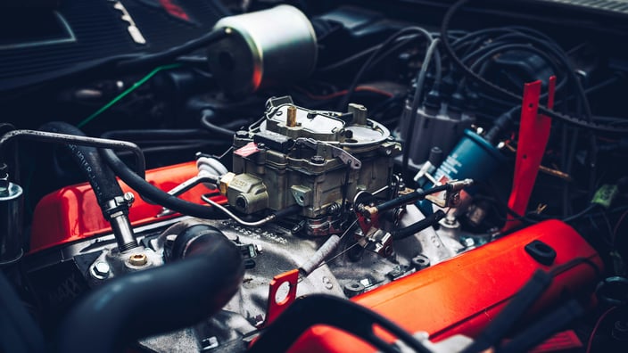 Detailní pohled na otevřený motor auta s červenými kryty ventilů a karburátorem v servisní dílně.