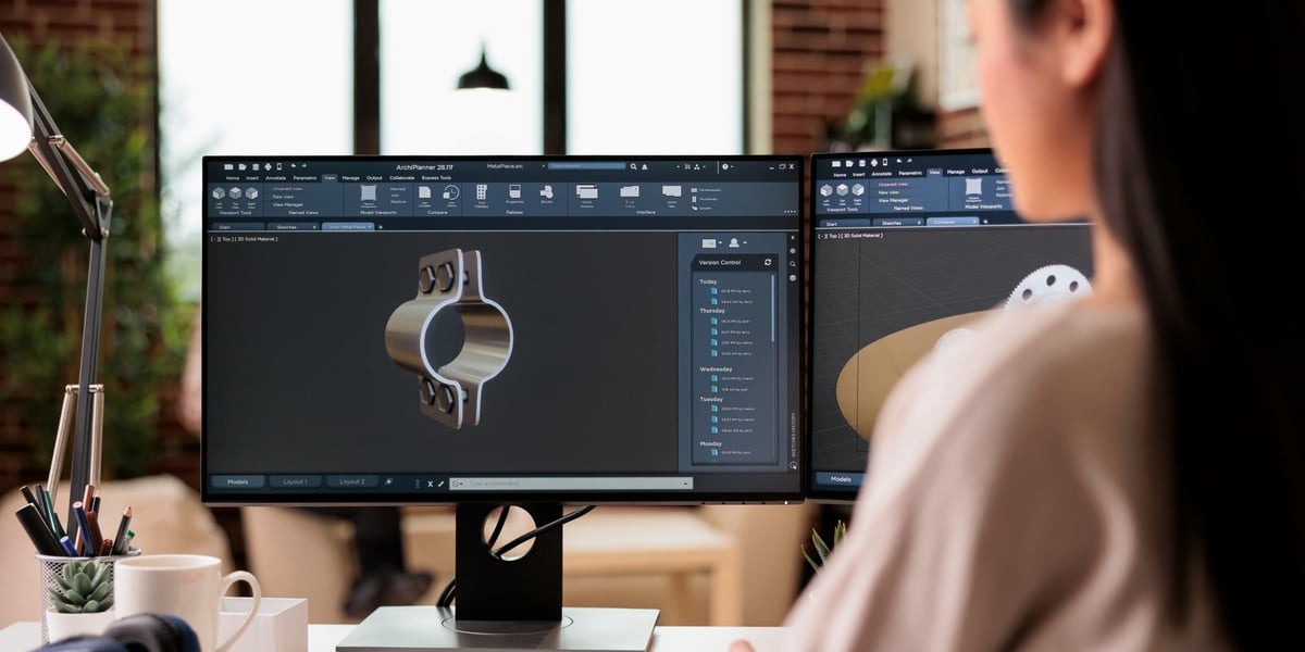 Pohled přes rameno na ženu, která sedí u stolu a pracuje na počítači se dvěma monitory. Na hlavní obrazovce je otevřený software pro 3D modelování (CAD), ve kterém navrhuje kovovou mechanickou součástku ve tvaru objímky. V pozadí je vidět moderní interiér s cihlovou zdí a velkým oknem.