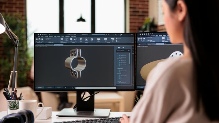 Pohled přes rameno na ženu, která sedí u stolu a pracuje na počítači se dvěma monitory. Na hlavní obrazovce je otevřený software pro 3D modelování (CAD), ve kterém navrhuje kovovou mechanickou součástku ve tvaru objímky. V pozadí je vidět moderní interiér s cihlovou zdí a velkým oknem.