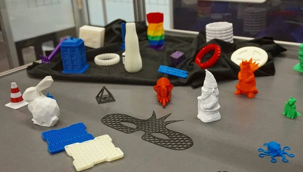 Výstava různých 3D vytištěných objektů, včetně bílého králíka, žlutého a modrého plástvového vzoru a černé masky uprostřed. Mezi dalšími předměty jsou barevné a bílé vázy a náramky.