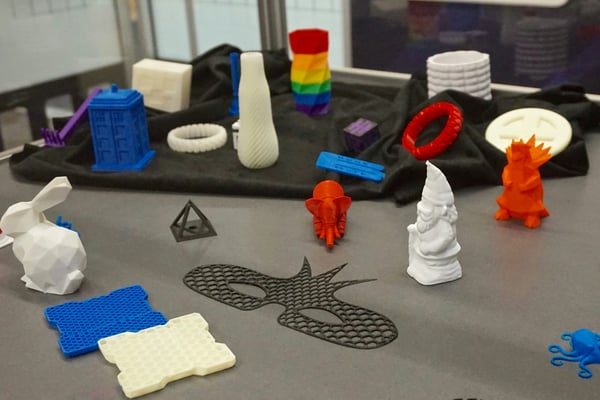 Výstava různých 3D vytištěných objektů, včetně bílého králíka, žlutého a modrého plástvového vzoru a černé masky uprostřed. Mezi dalšími předměty jsou barevné a bílé vázy a náramky.