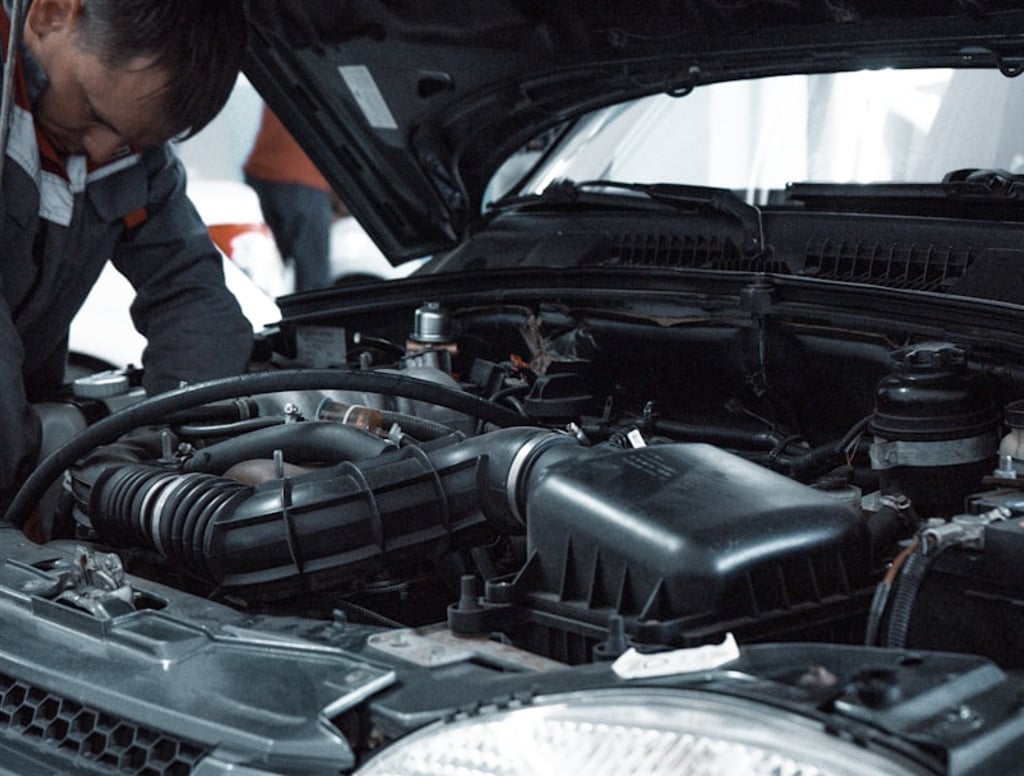 Mechanik v šedém pracovním oblečení se sklání nad otevřeným motorovým prostorem moderního vozu v dílně a kontroluje motor. V popředí je skříň vzduchového filtru a sací potrubí.