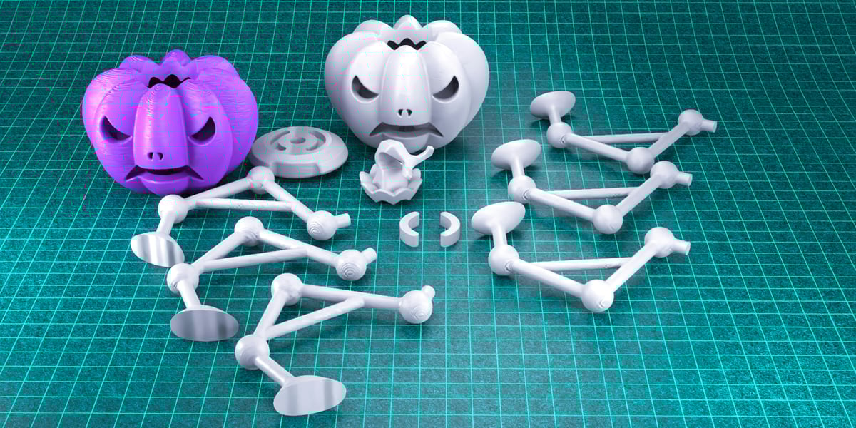 Zátiší zobrazující různé 3D tištěné předměty na tyrkysové řezací podložce s rastrem. Předměty zahrnují dvě figurky ve tvaru dýně (jedna fialová, jedna bílá/šedá) a několik bílých/šedých propojených modulárních plastových dílů kolem nich.