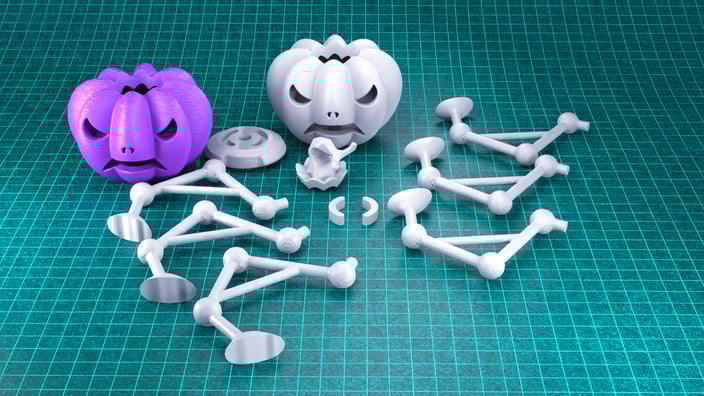 Zátiší zobrazující různé 3D tištěné předměty na tyrkysové řezací podložce s rastrem. Předměty zahrnují dvě figurky ve tvaru dýně (jedna fialová, jedna bílá/šedá) a několik bílých/šedých propojených modulárních plastových dílů kolem nich.