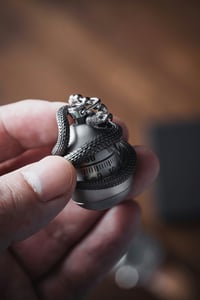 Detailní pohled na ruku držící elegantní 3D tištěný přívěsek se složitým motivem hada, který se obtáčí kolem kulaté kovové schránky.