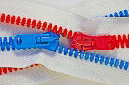 Dvě protikladné, červené a modré, plastové přezky od zipů se setkávají a spojují na bílém textilu. Znázorňuje výrobu zakázkových komponentů, jako jsou zipy, pro sportovní vybavení a oblečení.