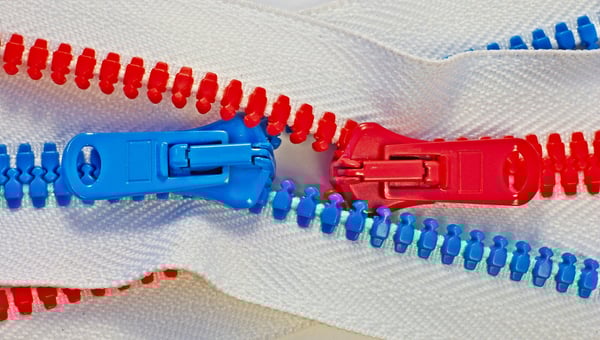 Dvě protikladné, červené a modré, plastové přezky od zipů se setkávají a spojují na bílém textilu. Znázorňuje výrobu zakázkových komponentů, jako jsou zipy, pro sportovní vybavení a oblečení.
