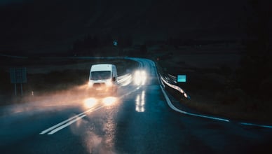 Вид из автомобиля на белый фургон, едущий по мокрой извилистой дороге ночью или в сумерках. Яркие фары фургона отражаются на влажном асфальте. На заднем плане — тёмные горы, окутанные густым туманом, что создаёт мрачную и атмосферную сцену.