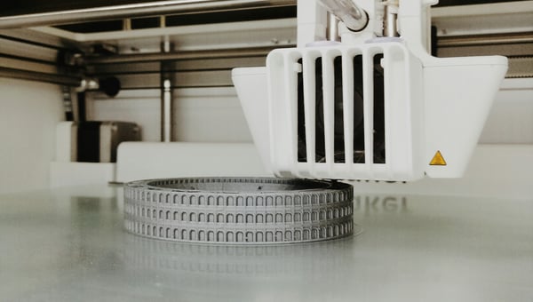 3D tiskárna vytváří detailní model v šedé barvě, který připomíná oblouky starověkého Kolosea. Ilustrace použití 3D tisku pro obnovu historických památek a artefaktů.