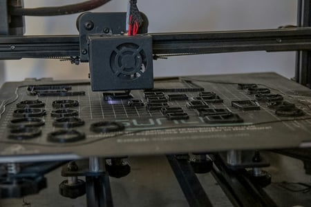 3D tiskárna s tryskou, která právě tiskne několik malých, černých kruhových dílů na tiskové podložce. Ilustruje výrobu zakázkových komponent a malých sérií.