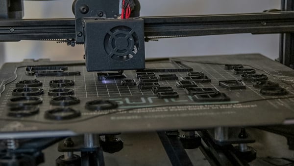 3D tiskárna s tryskou, která právě tiskne několik malých, černých kruhových dílů na tiskové podložce. Ilustruje výrobu zakázkových komponent a malých sérií.