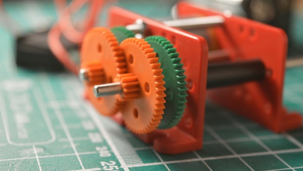 Detailní pohled na 3D vytištěný prototyp robotických dílů, včetně červených a zelených ozubených kol, umístěných na zelené podložce s mřížkou. Ilustruje výrobu komponent pro robotiku a automatizaci.