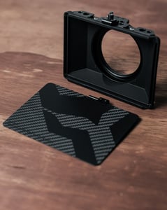 Černá matná 3D vytištěná sluneční clona a boční panel s texturou uhlíkových vláken. Ilustruje výrobu zakázkového a lehkého vybavení pro fotoaparáty a optiku.