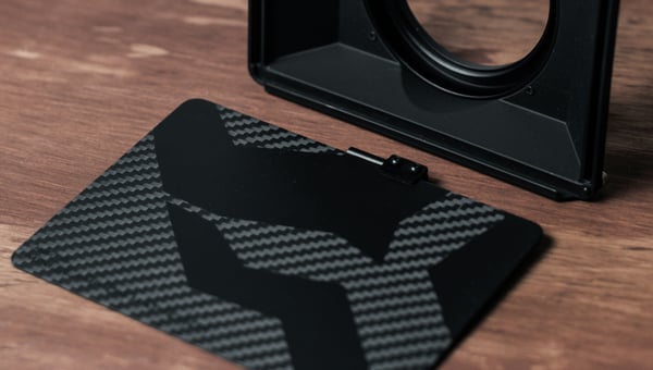 Černá matná 3D vytištěná sluneční clona a boční panel s texturou uhlíkových vláken. Ilustruje výrobu zakázkového a lehkého vybavení pro fotoaparáty a optiku.