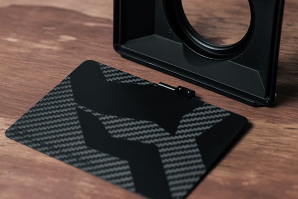 Černá matná 3D vytištěná sluneční clona a boční panel s texturou uhlíkových vláken. Ilustruje výrobu zakázkového a lehkého vybavení pro fotoaparáty a optiku.