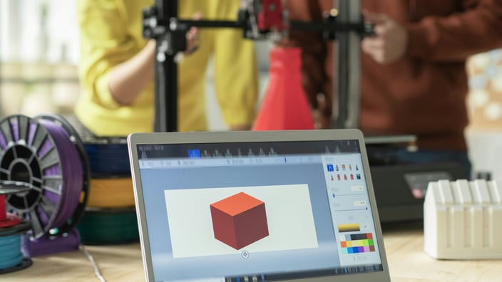 "Laptop s 3D modelem na stole v popředí, za ním 3D tiskárna s lidmi, kteří se na ni dívají