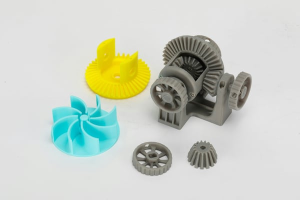 Skupina 3D vytištěných mechanických dílů různých barev a tvarů, včetně žluté vrtule, tyrkysového rotoru a šedých ozubených kol. Tyto díly ilustrují možnosti výroby náhradních a zakázkových komponent pro lodě a vodní plavidla.