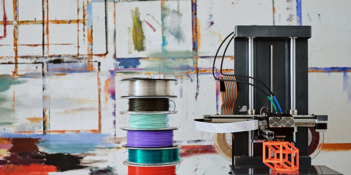 Moderní černá 3D tiskárna stojí vedle vysokého komínu cívek s filamentem v různých barvách (stříbrná, černá, modrozelená, fialová, červená, zelená) na bílém podkladu, před abstraktním malovaným pozadím. Na podložce tiskárny je malý, oranžový, geometrický zkušební výtisk.