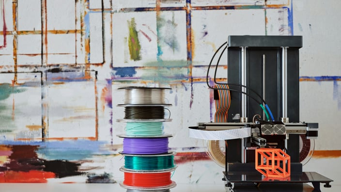 Moderní černá 3D tiskárna stojí vedle vysokého komínu cívek s filamentem v různých barvách (stříbrná, černá, modrozelená, fialová, červená, zelená) na bílém podkladu, před abstraktním malovaným pozadím. Na podložce tiskárny je malý, oranžový, geometrický zkušební výtisk.