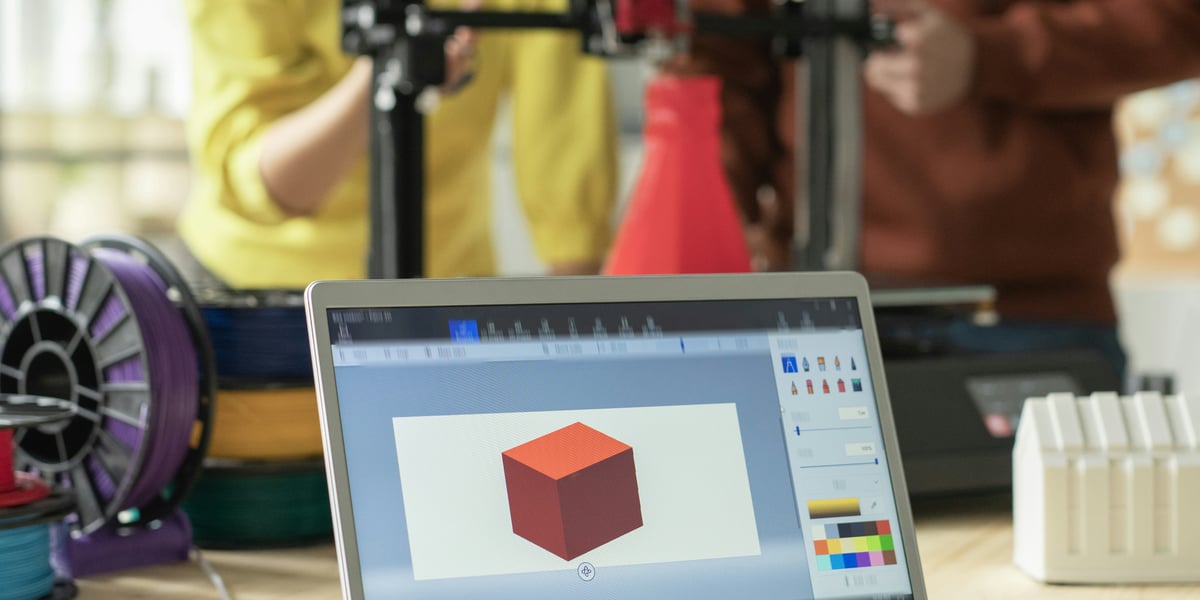 3Studenti a učitelé se učí pracovat s 3D tiskárnou ve třídě, s notebookem zobrazujícím 3D model krychle v popředí, symbolizující STEM vzdělávání.