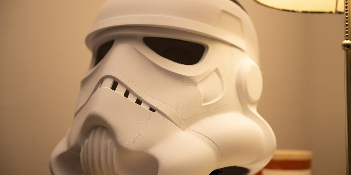 A 3D printed Stormtrooper helmet, in an unfinished matte white state with visible layer lines from the printing process, rests on a wooden surface next to a lamp, showcasing a work-in-progress cosplay prop.