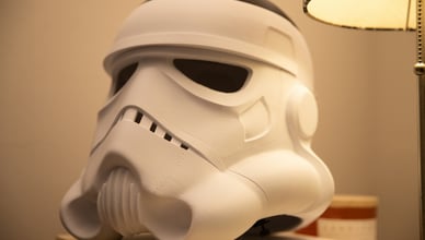A 3D printed Stormtrooper helmet, in an unfinished matte white state with visible layer lines from the printing process, rests on a wooden surface next to a lamp, showcasing a work-in-progress cosplay prop.