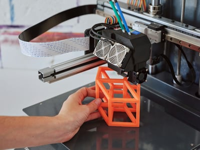 Detailní záběr na ruku, která odstraňuje oranžový, geometrický 3D vytištěný prototyp z tiskové podložky 3D tiskárny.