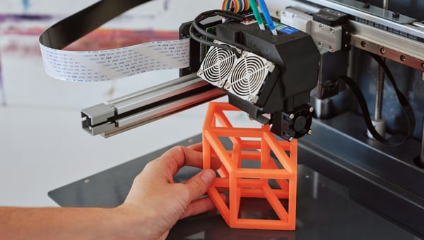 Detailní záběr na ruku, která odstraňuje oranžový, geometrický 3D vytištěný prototyp z tiskové podložky 3D tiskárny.