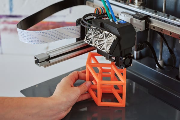 Detailní záběr na ruku, která odstraňuje oranžový, geometrický 3D vytištěný prototyp z tiskové podložky 3D tiskárny.