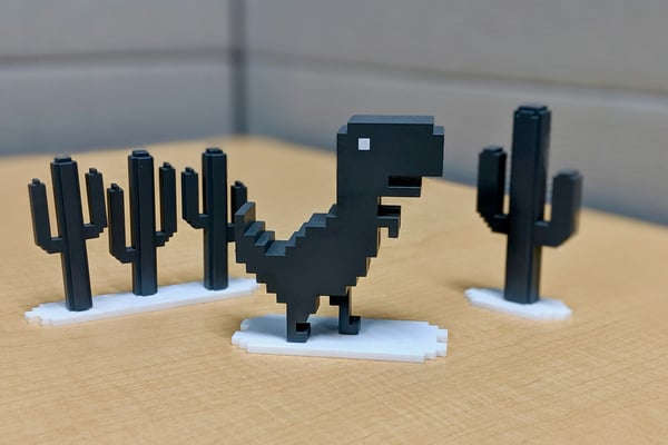 Malé, 3D vytištěné figurky v pixelové grafice na stole. Je na nich šedý dinosaurus a několik kaktusů, což odkazuje na populární online hru a ukazuje, jak lze 3D tiskem vytvářet vlastní figurky pro stolní hry.